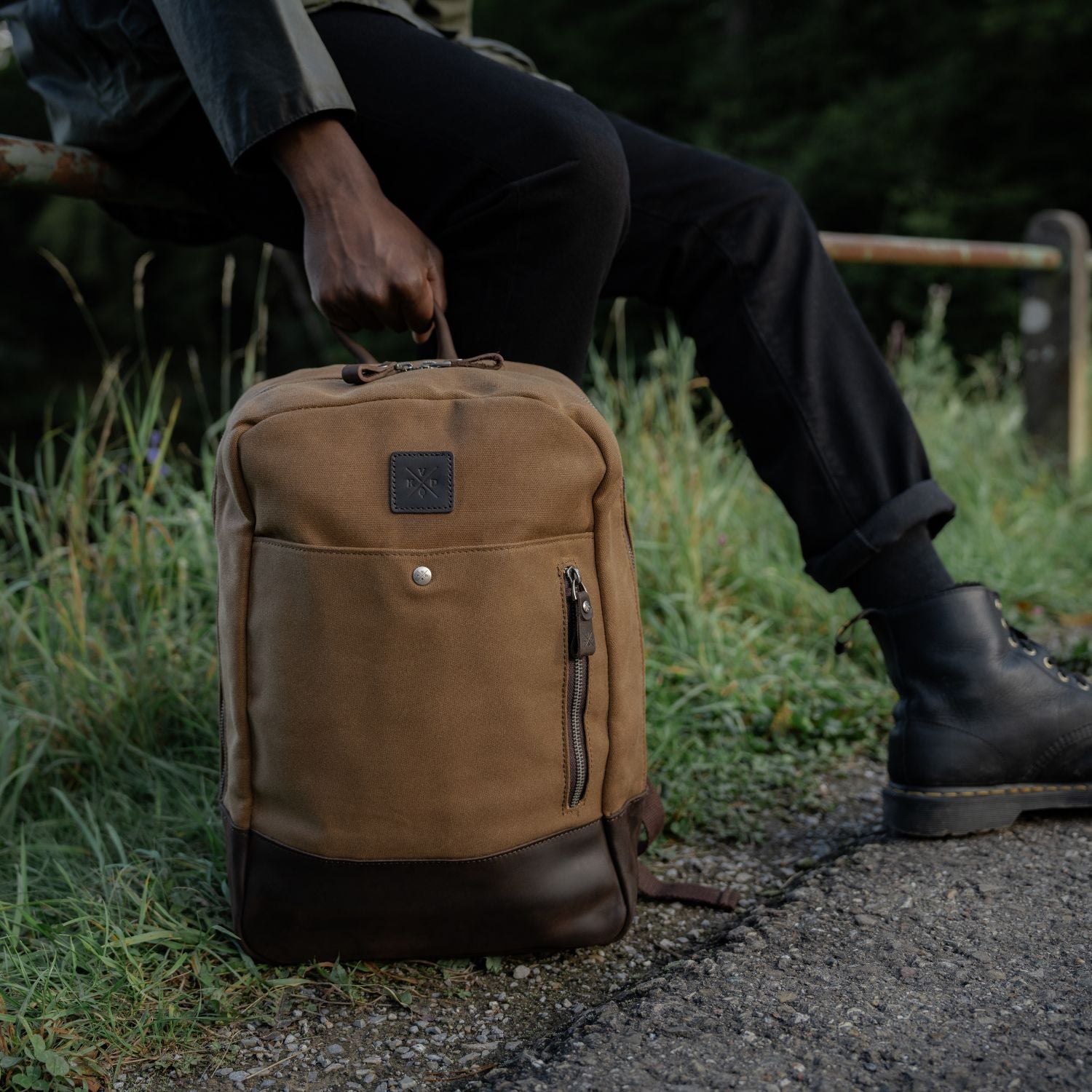 Tan waxed canvas and reclaimed leather Eden backpack on the floor being held by the leather carry handle