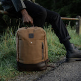 Tan waxed canvas and reclaimed leather Eden backpack on the floor being held by the leather carry handle