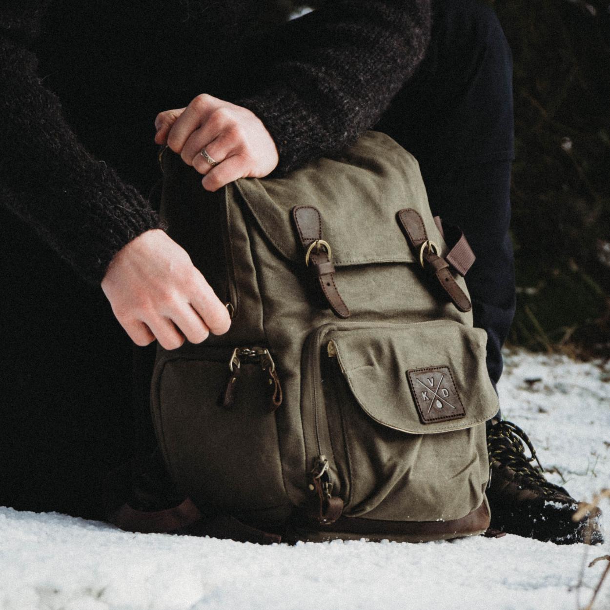 Man knelt in the snow unzipping one of the side pockets on the Roe camera bag/backpack