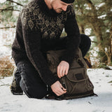 Man kneeling in the snow unzipping the camera pocket on the front of the Roe waxed canvas backpack