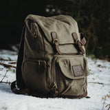 Roe - Moss Green waxed canvas backpack sat in the snow within a wooded area.