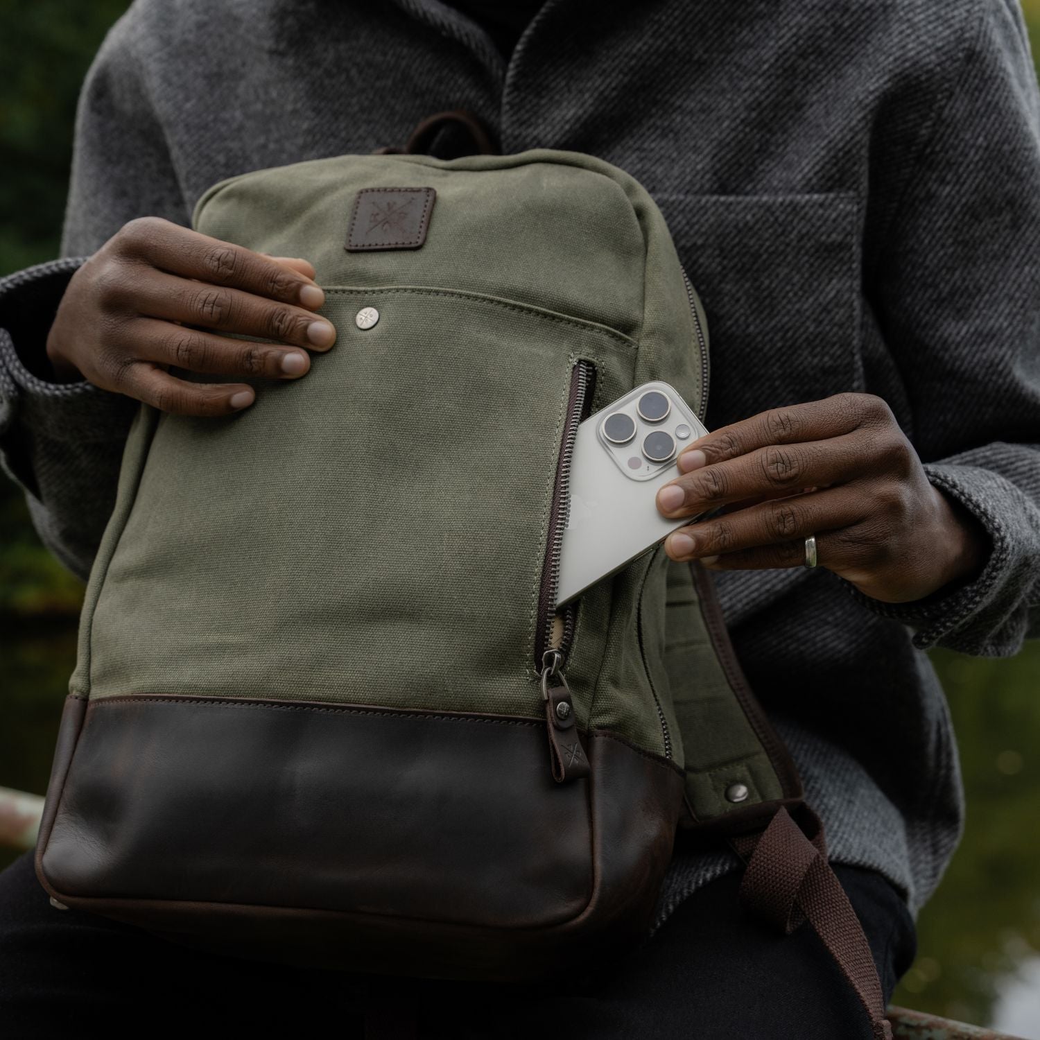 Man putting phone in front zipped pocket of Moss Green waxed canvas Eden backpack 