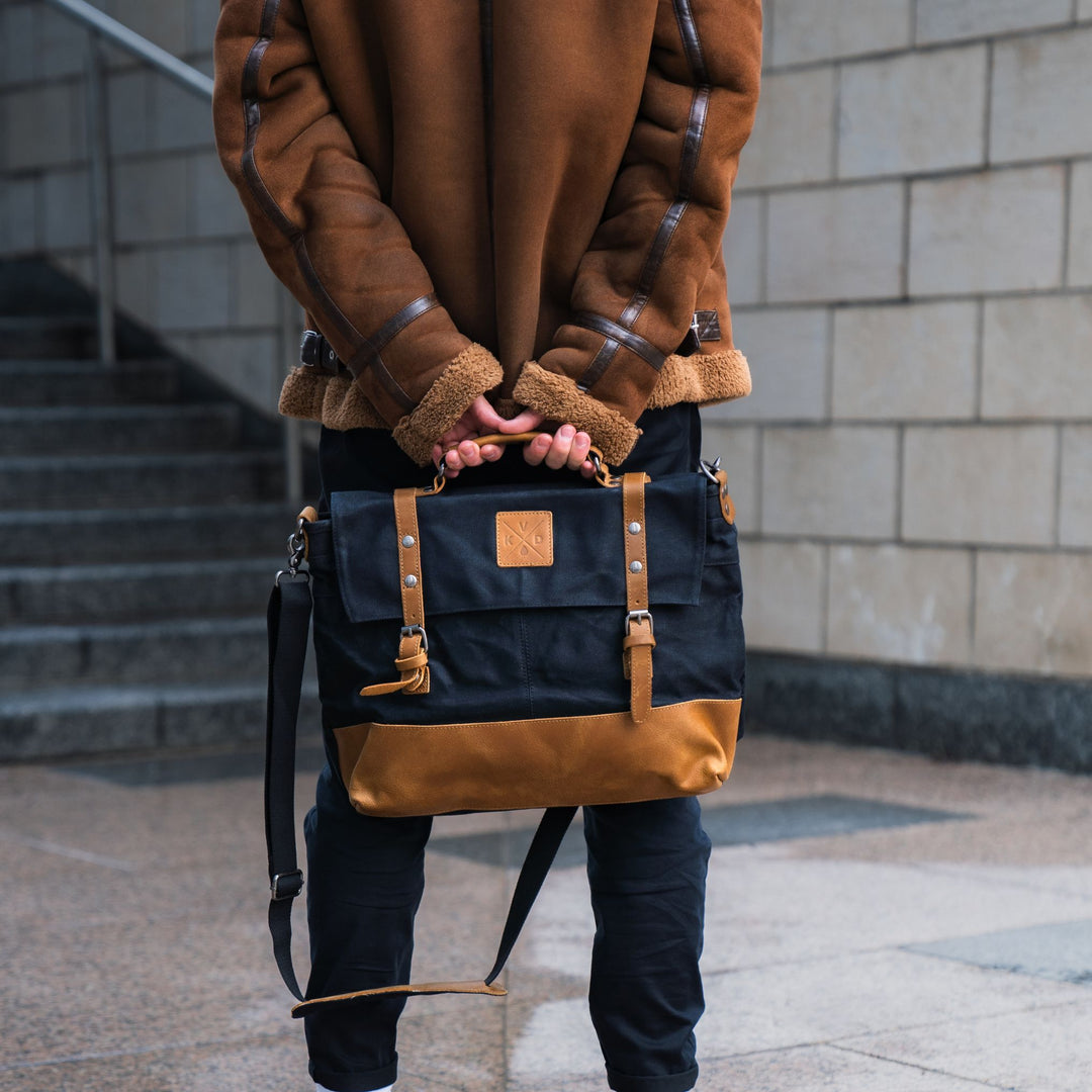 Mersey black messenger bag held by model in brown jacket#colour_black