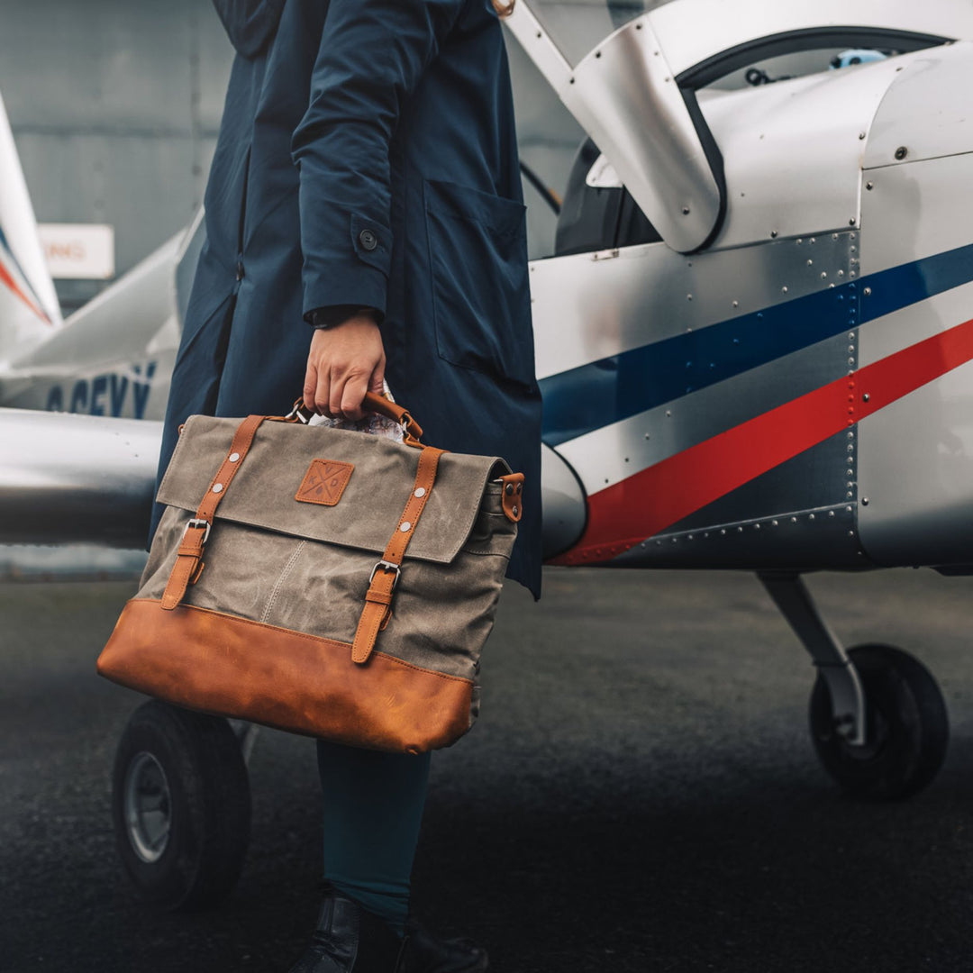 Kovered Mersey moss green messenger style satchel bag held by model walking in front of a plane#colour_moss-green
