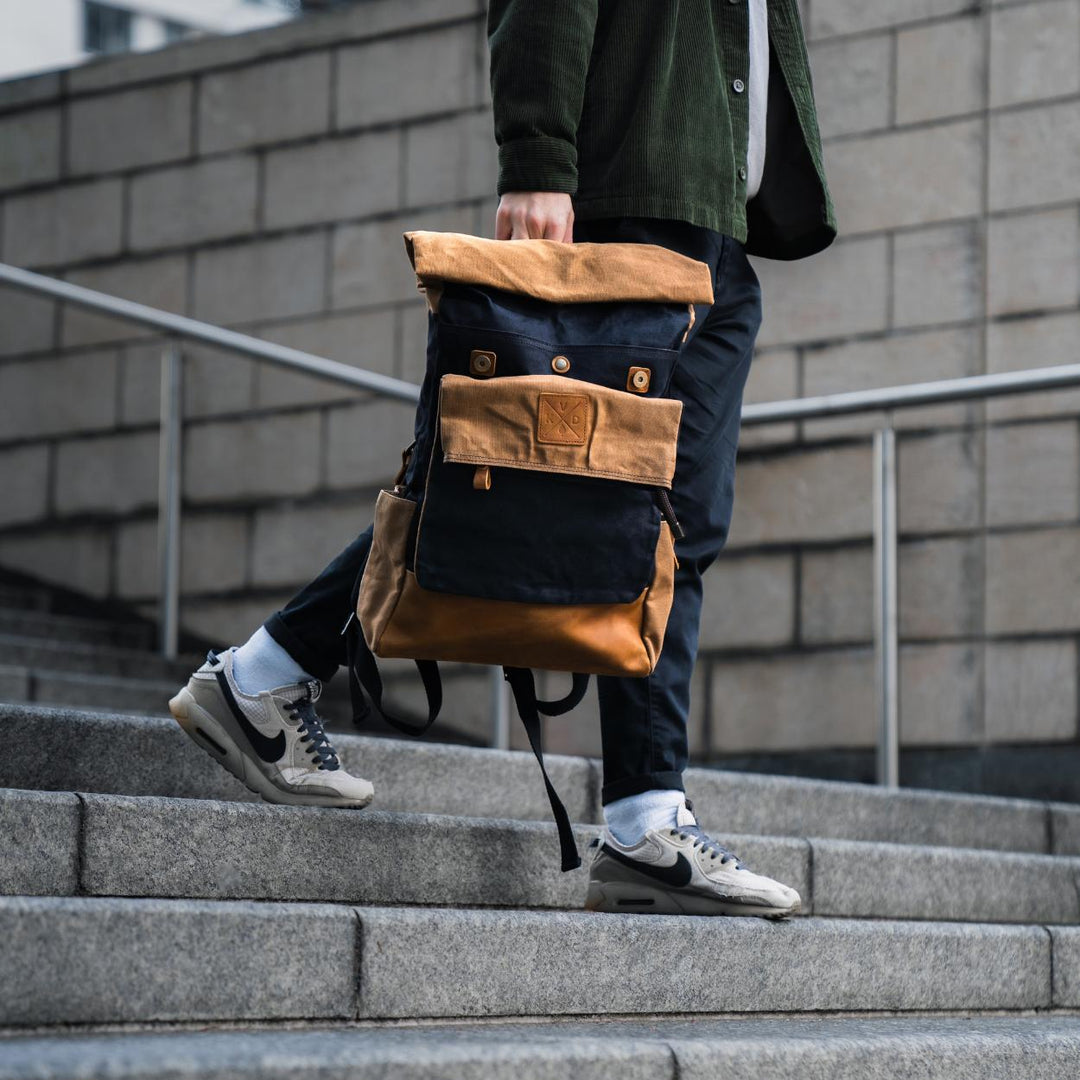 Kovered taw black and tan rolltop backpack held by model walking down some stairs#colour_black