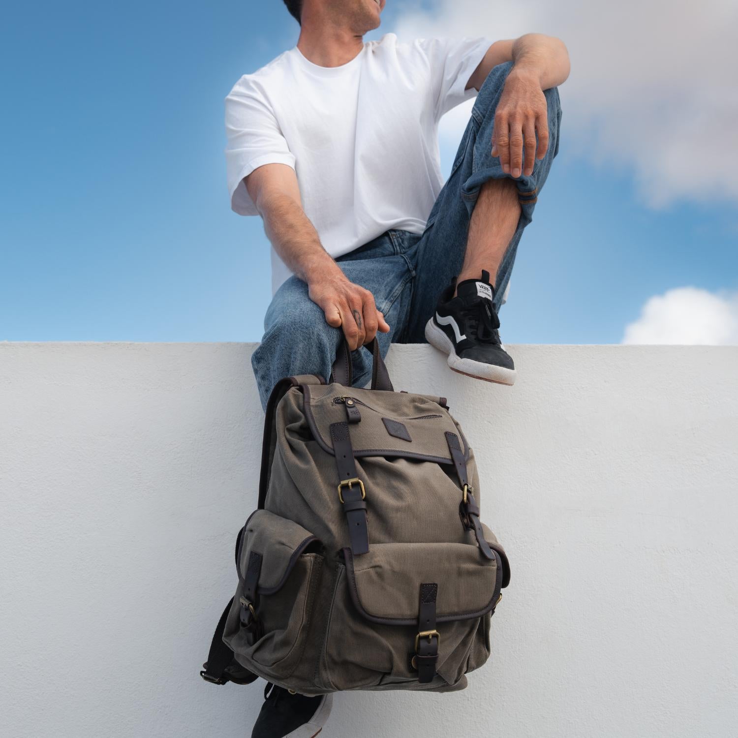 Man sat on a wall holding the Lune backpack with the top leather carry handle.