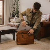 Man putting laptop in Manifold - Tan leather messenger bag with zipped front pocket and carry handles 