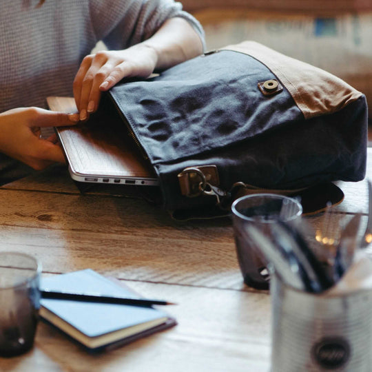Kovered Medway messenger bag on a coffee table with a model taking the laptop out of the laptop pocket#colour_navy