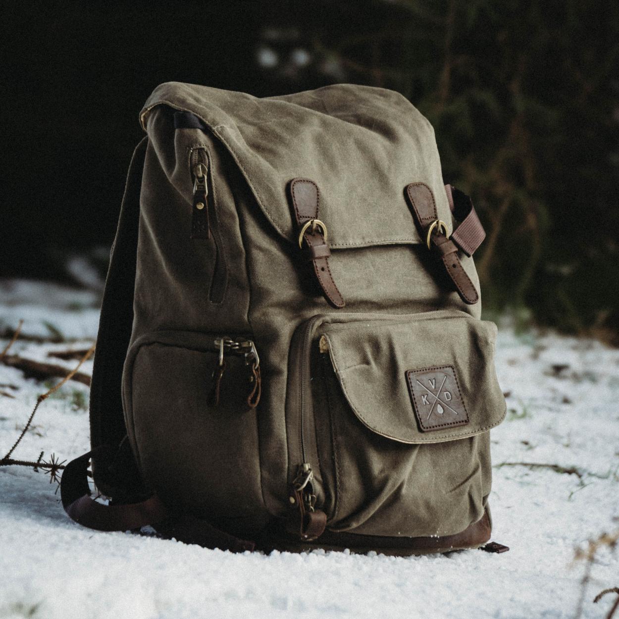 Roe - Moss Green waxed canvas backpack sat in the snow within a wooded area.